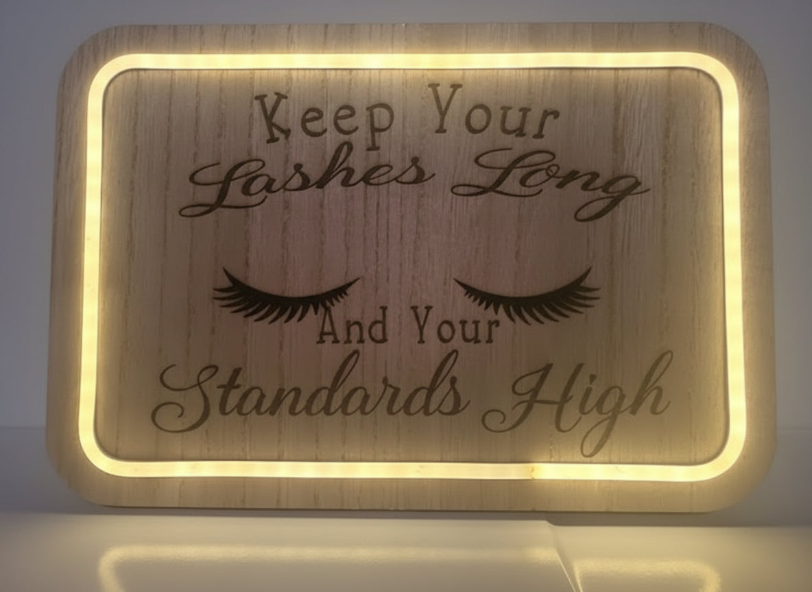 Wooden sign with engraved eyelashes and text 'Keep Your Lashes Long And Your Standards High' on a light background.