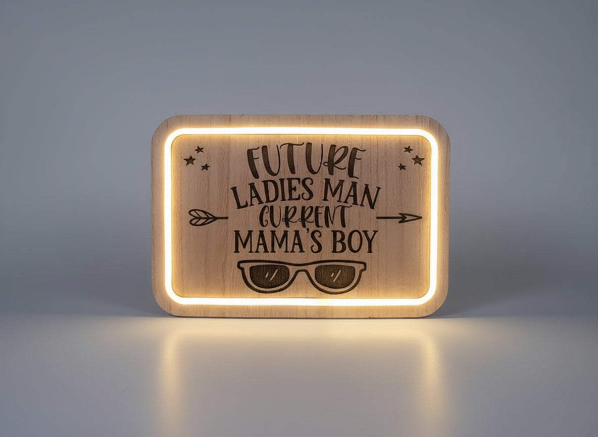 Wooden plaque with engraved text 'Future Ladies Man, Current Mama's Boy' and sunglasses design, illuminated from behind.