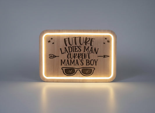 Wooden plaque with engraved text 'Future Ladies Man, Current Mama's Boy' and sunglasses design, illuminated from behind.