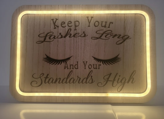Wooden sign with engraved eyelashes and text 'Keep Your Lashes Long And Your Standards High' on a light background.
